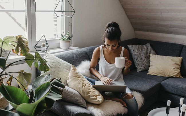 Jeune femme assise sur le canapé avec une tasse de café à l'aide d'un ordinateur portable