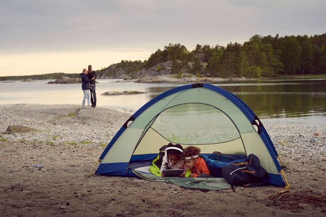 Frères et sœurs allongés avec une tablette numérique dans une tente tandis que les parents se tiennent à la plage pendant le coucher du soleil
