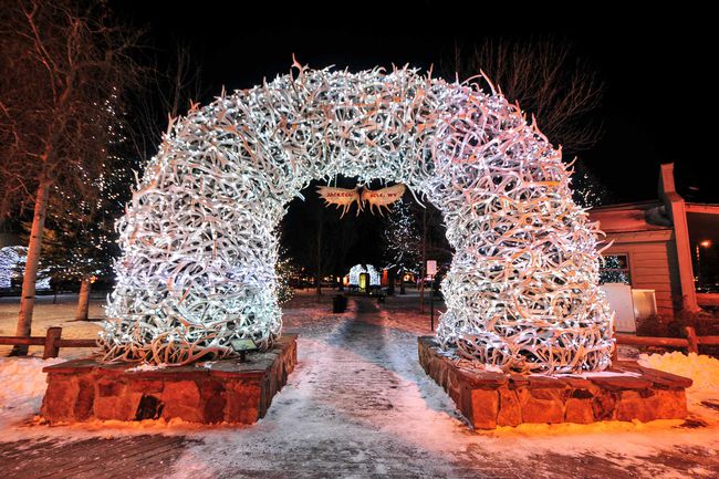 De grandes arches en bois de wapiti se courbent au-dessus des quatre entrées d'angle de la place de Jackson Hole.  Les bois sont là depuis le début des années 1960, et de nouveaux arcs sont actuellement assemblés pour les remplacer.  Illuminé la nuit pour Noël.