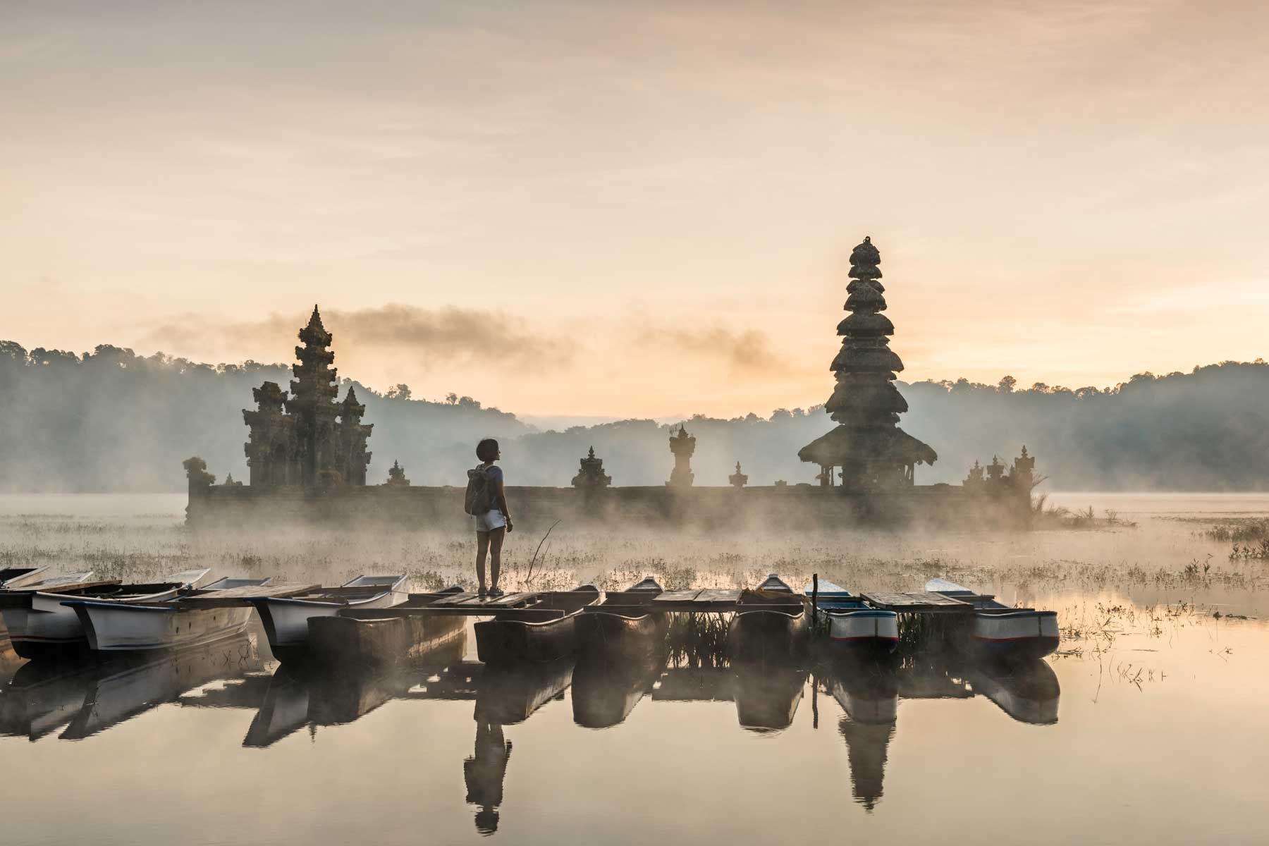 Jeune femme asiatique debout sur un bateau admirant le temple de Tamblingan, immergé dans l'eau, avec une brume matinale enveloppant le temple hindou.