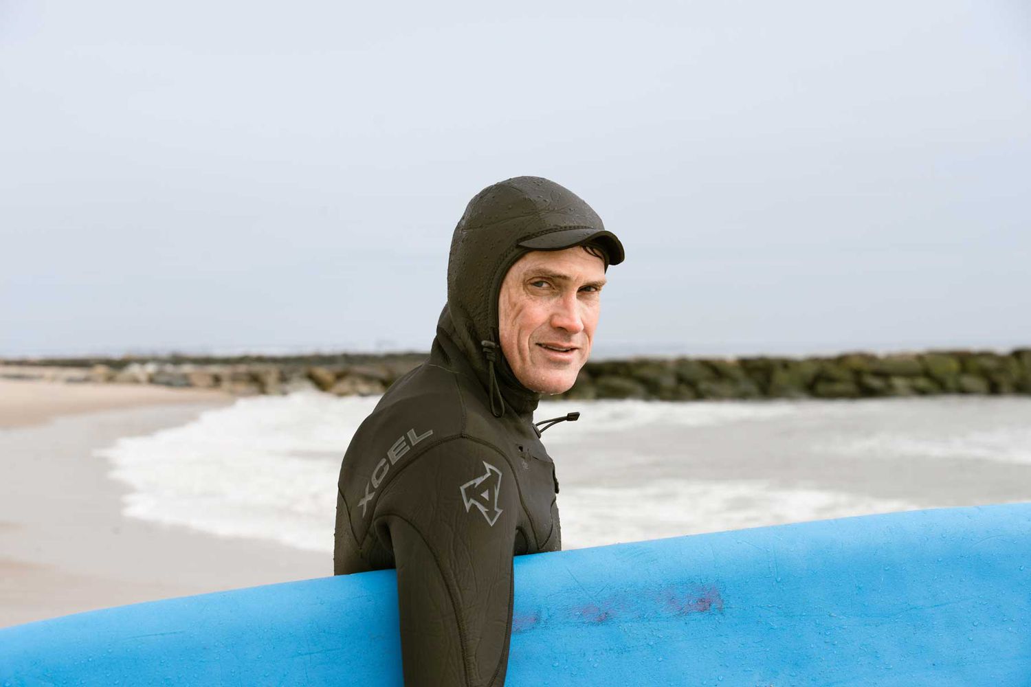 L'écrivain Tom Vanderbilt portant une combinaison intégrale et tenant une planche de surf bleue