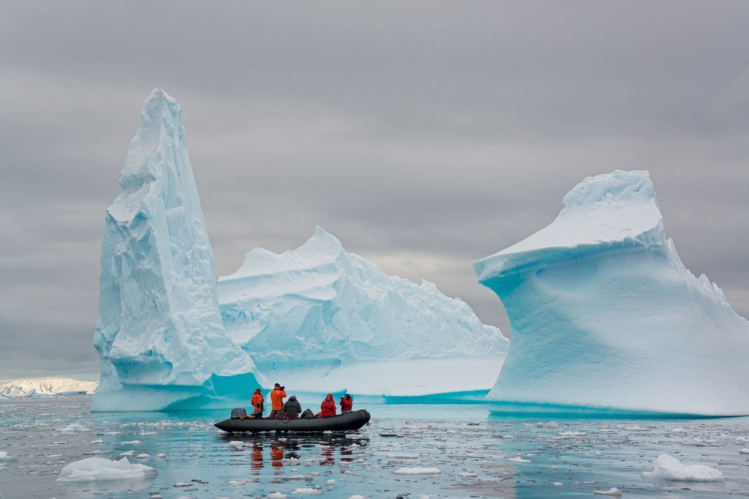 Des gens dans de petits bateaux à côtes gonflables en zodiac passant d'imposants icebergs sculptés sur l'eau calme autour de petites îles de la péninsule antarctique.