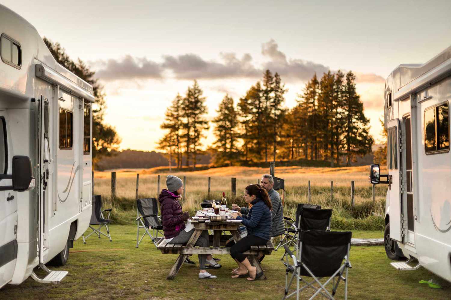 Homme et deux femmes en train de dîner à l'extérieur autour d'une table de pique-nique en bois au coucher du soleil